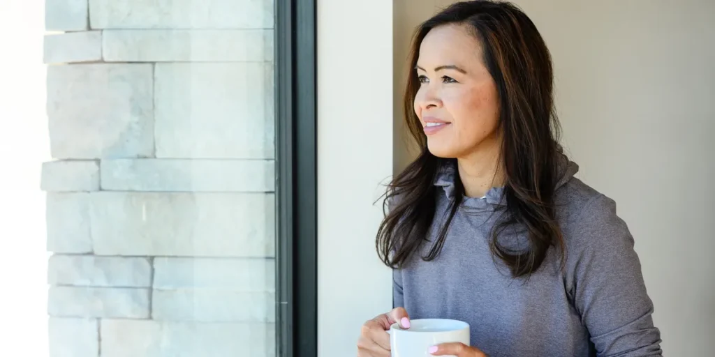 Dr. Angela Tran, DO holding a mug and looking out a window at home
